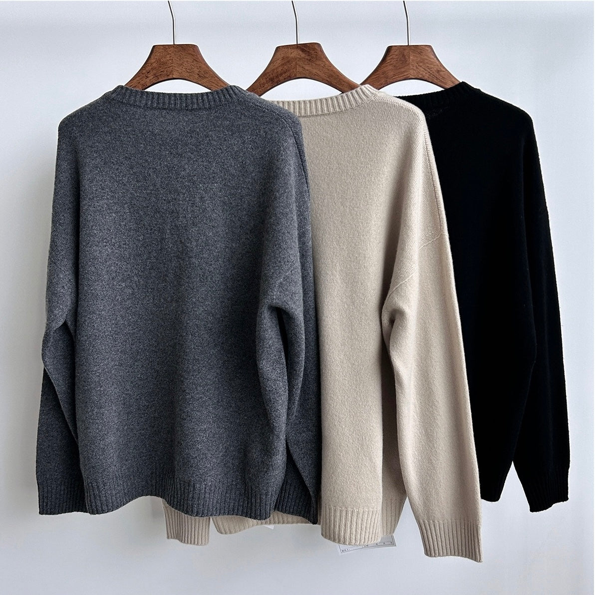 Three sweaters in gray, beige, and black hanging on wooden hangers against a white background.