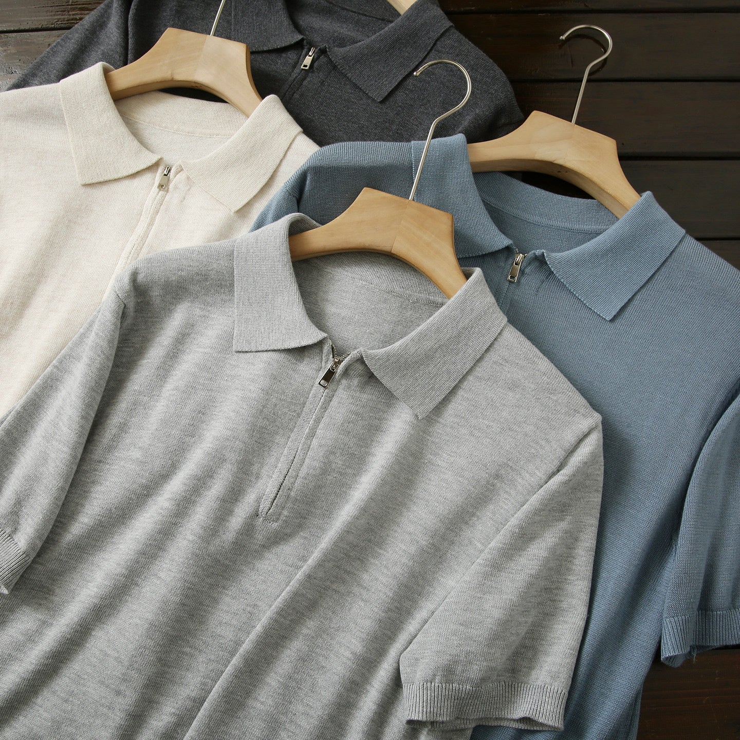 Four polo shirts in beige, gray, blue, and black on hangers against a wooden background.